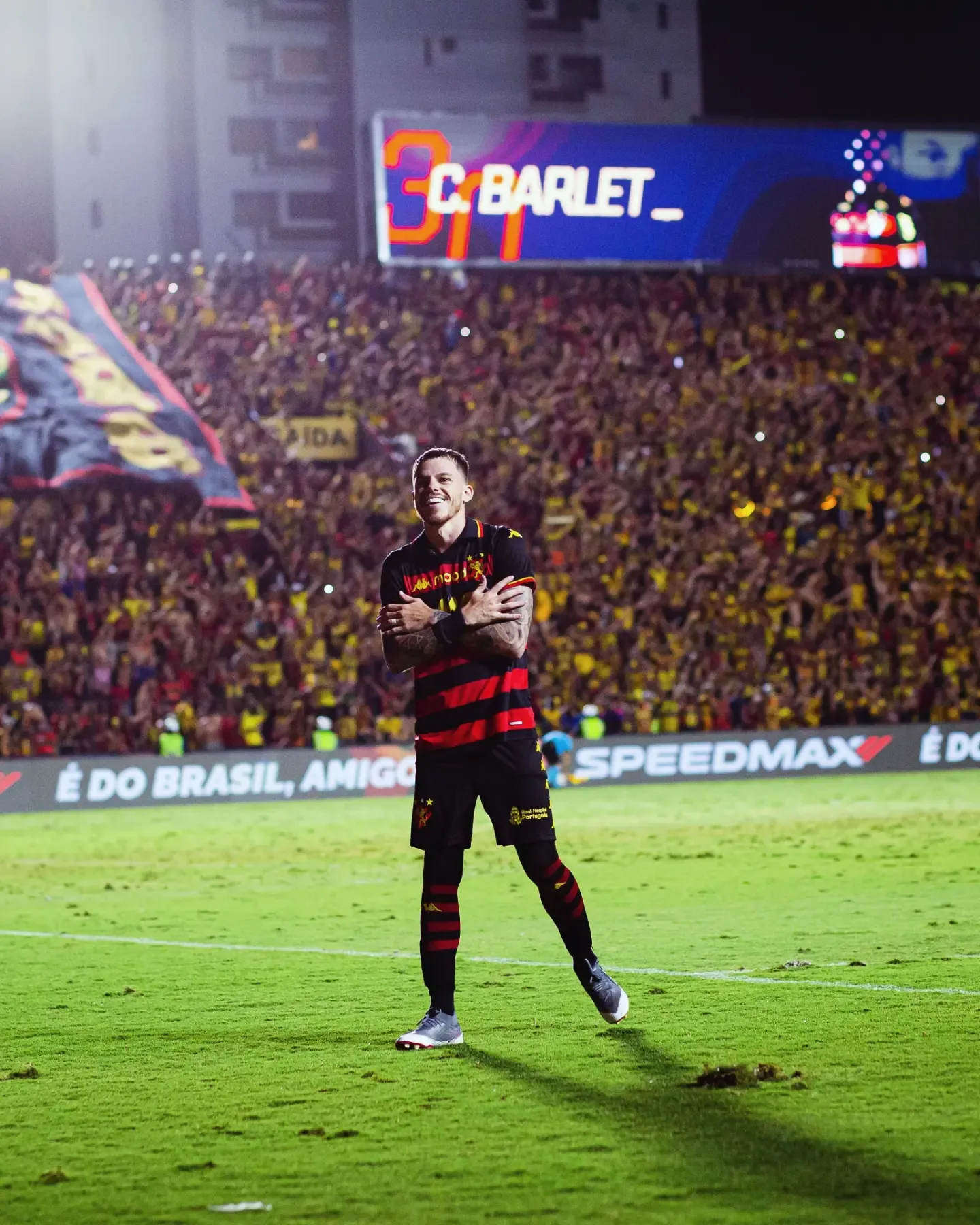 Jogador de uniforme preto e vermelho, de braços cruzados sobre o peito, sorrindo em frente à arquibancada lotada de um estádio.