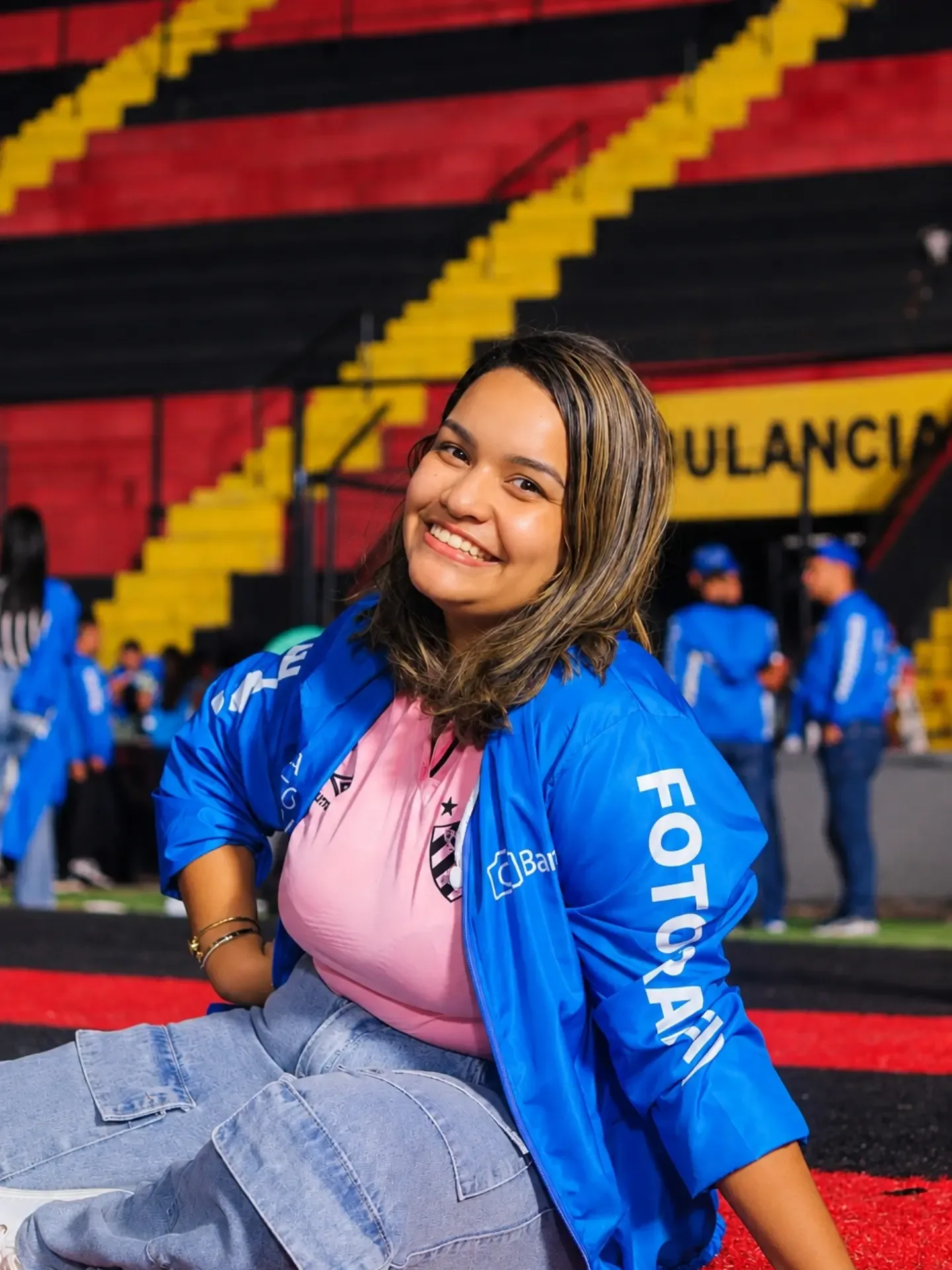 Hellen Maria sentada no gramado de um estádio, sorrindo para a câmera, com jaqueta azul e arquibancada vermelha e preta ao fundo.