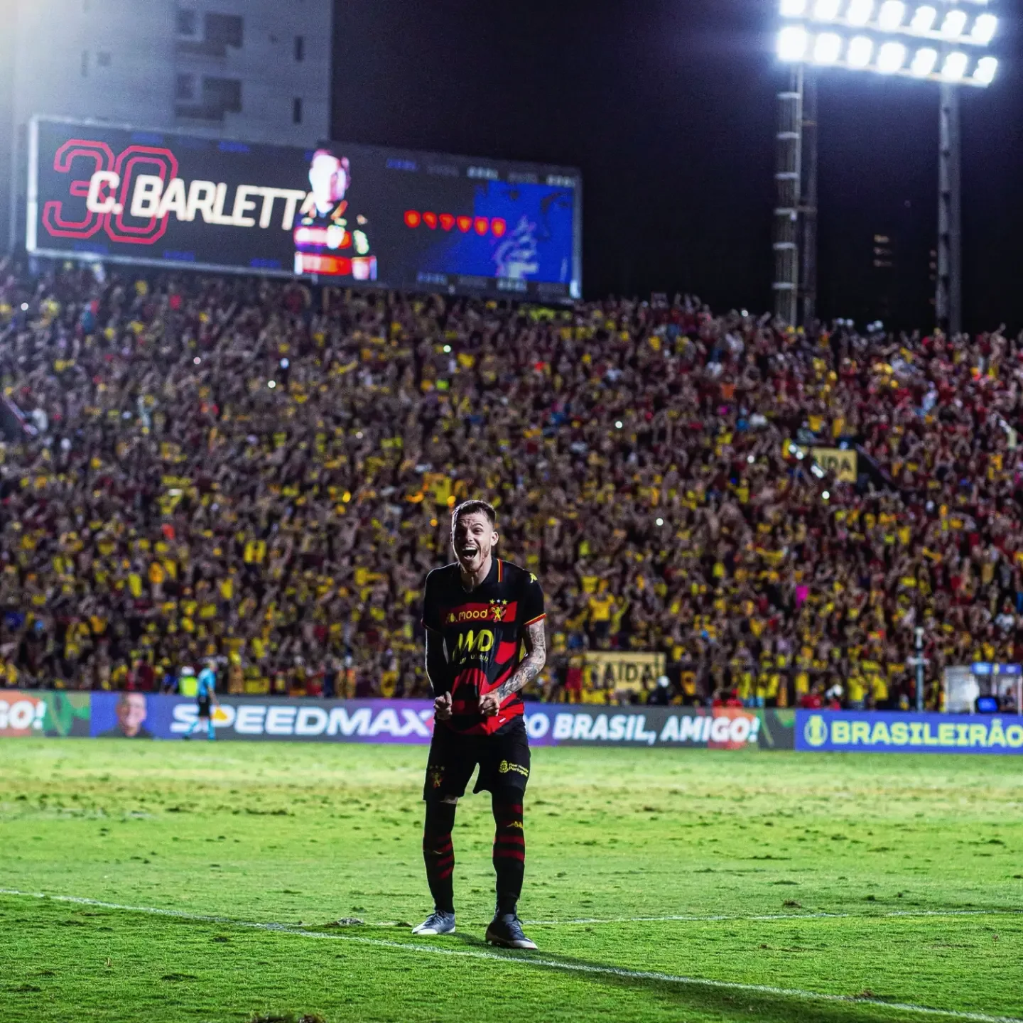 Jogador de uniforme preto e vermelho comemorando aos gritos, com os punhos fechados, no gramado de um estádio à noite, com a torcida e o placar atrás.