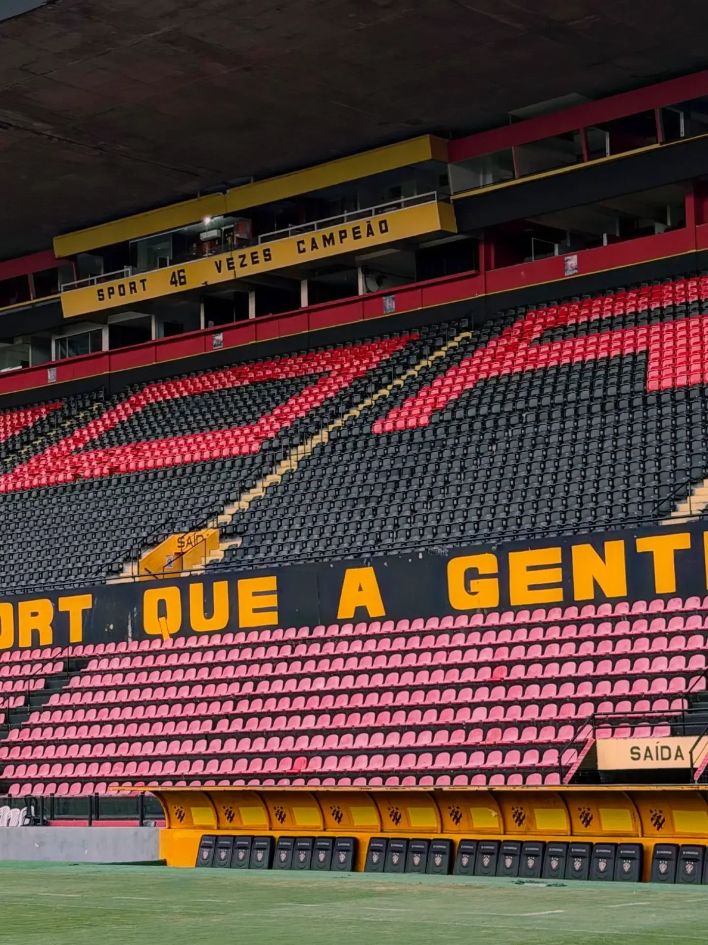 Arquibancada de um estádio com cadeiras pretas e vermelhas, uma faixa preta e amarela e a cabine de imprensa ao alto.