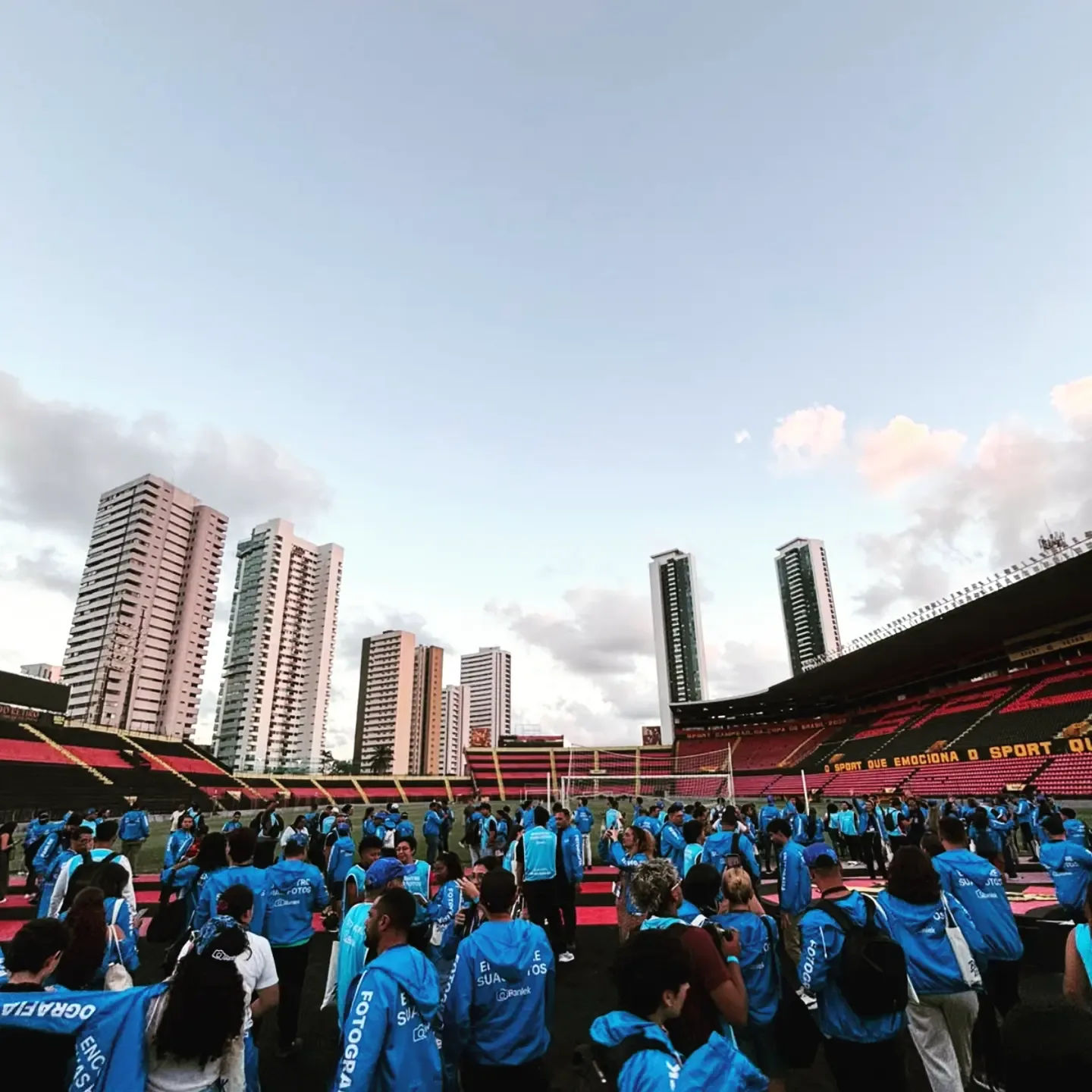 Fotógrafos de colete azul reunidos no gramado de um estádio ao entardecer, com prédios e nuvens ao fundo.