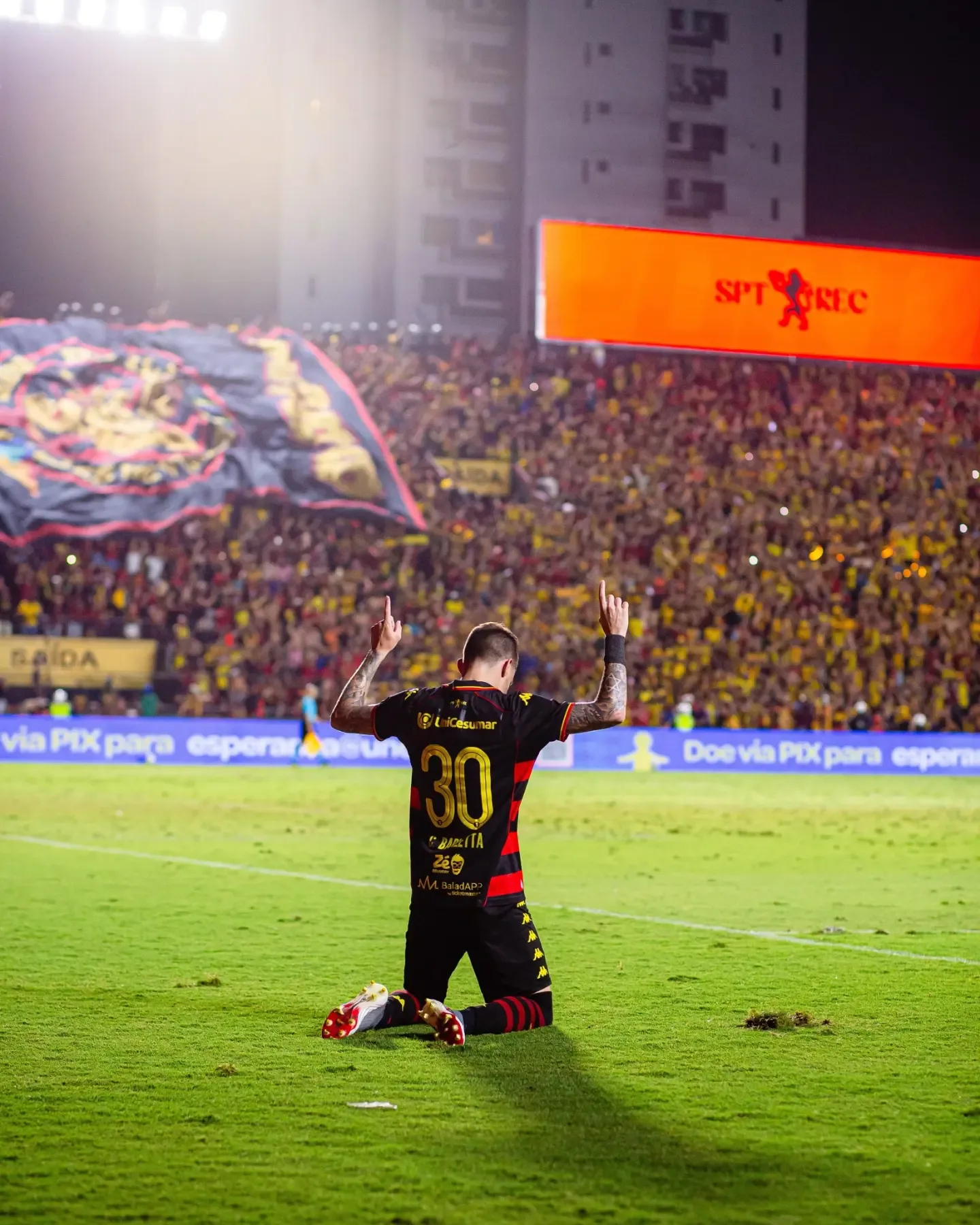 Jogador de uniforme preto e vermelho, de costas, ajoelhado no gramado com os braços erguidos diante da torcida de um estádio à noite.